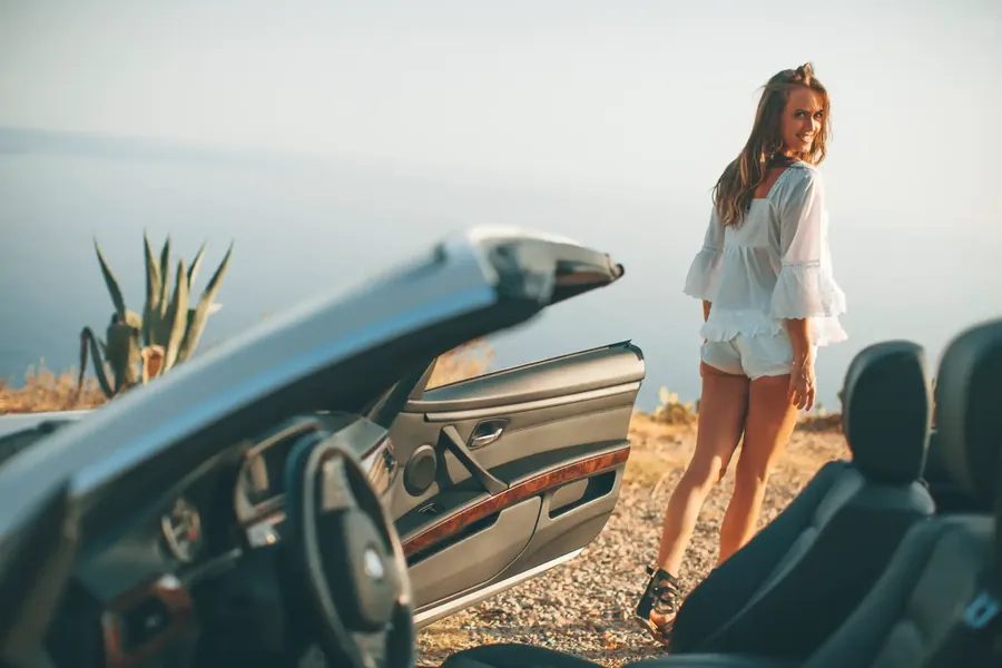 A woman in a straw hat beside a convertible car overlooking the Pacific