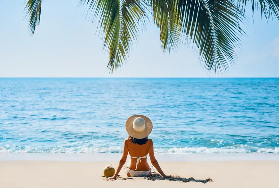 Palmiana from behind, in a straw hat on a tropical beach under a palm frond